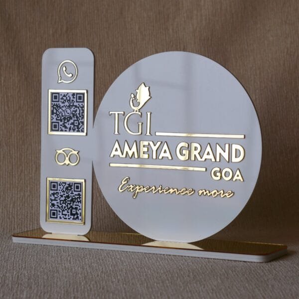 A custom white and gold acrylic tabletop sign for "TGI Ameya Grand Goa" with QR codes for WhatsApp and Google reviews, set on a reflective gold base.