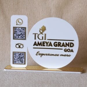 Front-facing view of a modern acrylic business sign with 3D gold lettering for "TGI Ameya Grand Goa" and two scannable QR codes for customer contact.