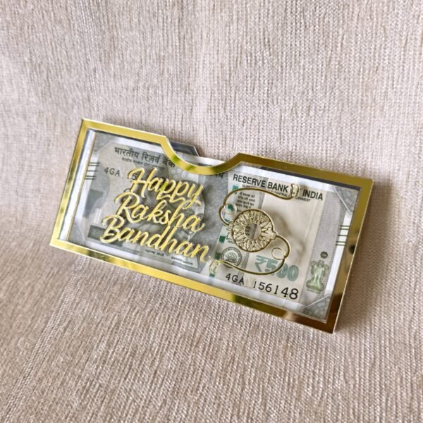 A stylish Raksha Bandhan money gift holder made of clear acrylic with shiny gold accents, with a 500 rupee note visible inside.