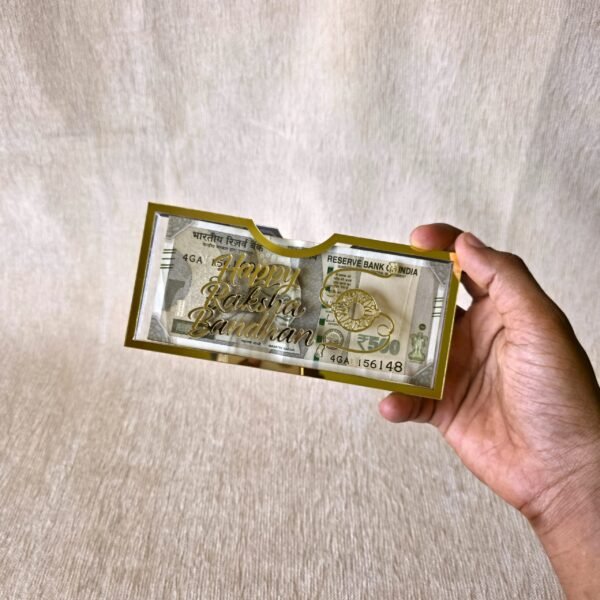 A hand holding the premium gold acrylic 'Happy Raksha Bandhan' money gift envelope to showcase its size and elegant design against a neutral background.