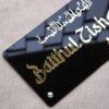 Angled view showing the 3D effect of the golden 'Baithul Zishaan' lettering on the glossy black acrylic nameplate.