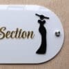 Close-up view of the black female silhouette icon in a gown and a silver mounting stud on the 'Female Section' wall sign.