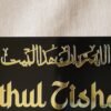 Close-up of the golden Arabic calligraphy 'Allahumma barik hazal bait' on a black acrylic Islamic name plate.