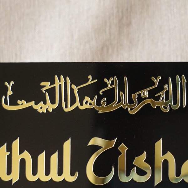 Close-up of the golden Arabic calligraphy 'Allahumma barik hazal bait' on a black acrylic Islamic name plate.