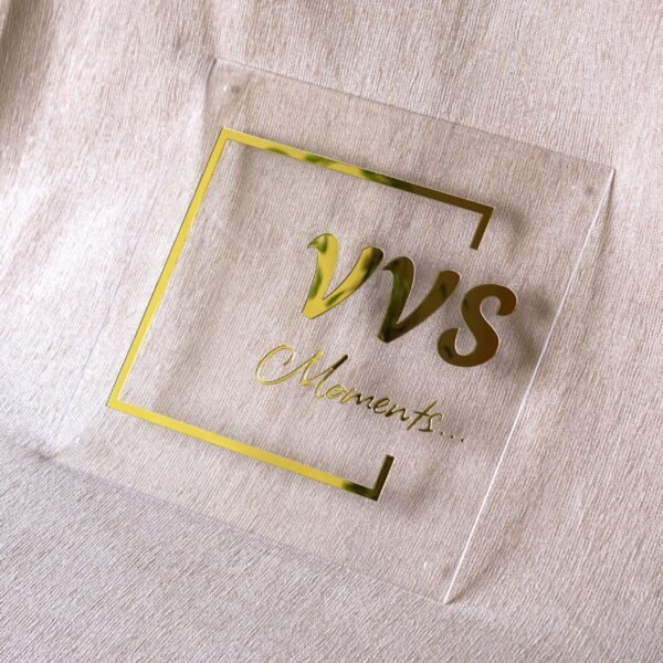 Front view of a custom gold and clear acrylic business sign with the text "VVS Moments..." against a light-colored fabric background.