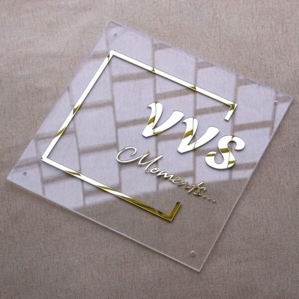 Angled side view of a square clear acrylic sign with 3D gold mirror lettering that reads "VVS Moments...", highlighting the raised text and reflections.