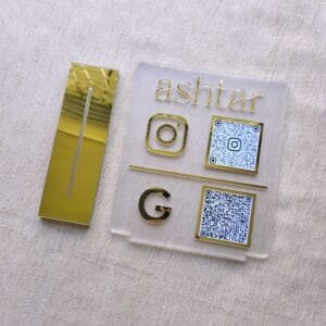 The custom QR code display stand disassembled, showing the gold mirror base next to the matte acrylic sign plate for 'ashtar'.