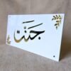 Angled view of a personalized Arabic nameplate with the name 'Jannat' in gold mirror calligraphy on a white acrylic base, showing its reflective surface.