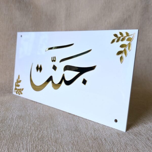 Angled view of a personalized Arabic nameplate with the name 'Jannat' in gold mirror calligraphy on a white acrylic base, showing its reflective surface.