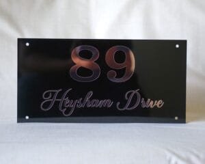 Front view of a custom 12x6 inch house sign showing "89 Heysham Drive" in rose gold lettering on a glossy black acrylic plate.