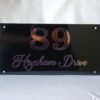 np2 Front view of a custom 12x6 inch house sign showing "89 Heysham Drive" in rose gold lettering on a glossy black acrylic plate.