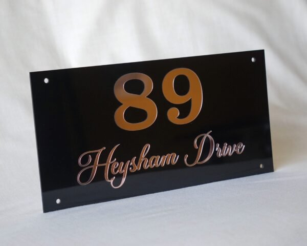 np4 Angled view of an elegant black acrylic nameplate for "89 Heysham Drive," highlighting the reflective quality of the rose gold custom text.