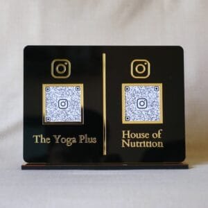 Front-facing view of a custom dual QR code display stand in black and gold acrylic, featuring Instagram links for 'The Yoga Plus' and 'House of Nutrition'.