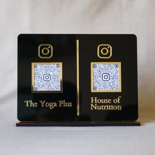 Front-facing view of a custom dual QR code display stand in black and gold acrylic, featuring Instagram links for 'The Yoga Plus' and 'House of Nutrition'.