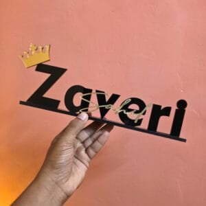 A hand holding a personalized black and gold acrylic desk nameplate with a crown against a salmon-pink wall.
