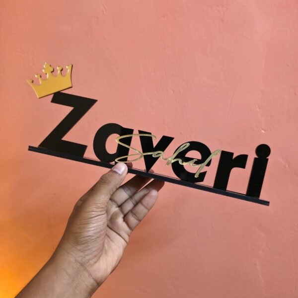 A hand holding a personalized black and gold acrylic desk nameplate with a crown against a salmon-pink wall.