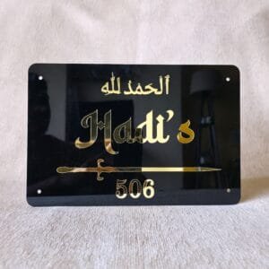 A straight-on view of a personalized Islamic nameplate in black acrylic, featuring a decorative sword and 'Alhamdulillah' in gold.