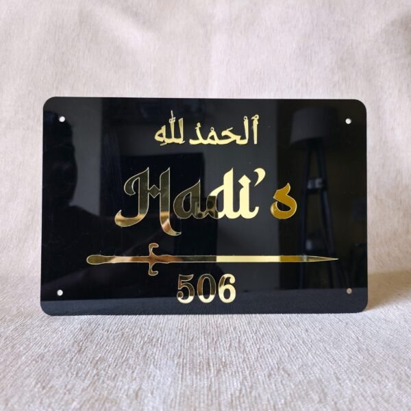 A straight-on view of a personalized Islamic nameplate in black acrylic, featuring a decorative sword and 'Alhamdulillah' in gold.