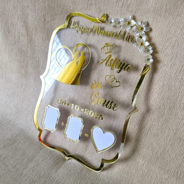 Tilted view of a transparent and gold acrylic wedding thumb board, personalized with names 'Asfiya' and 'Gouse' and a wedding date, showing reflections on its glossy surface against a light beige background.