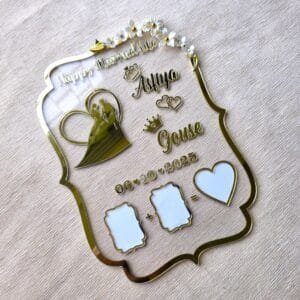 Angled top-down view of a personalized gold acrylic wedding thumb board with 'Asfiya' and 'Gouse' names, wedding date, and white floral accents, highlighting the transparent and reflective elements on a textured beige fabric.