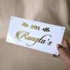 A hand holding up a white acrylic name plate with shiny 3D golden letters displaying "201 Rungta's" and decorative leaf motifs, against a textured beige background.