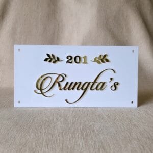 20250921_163216 A front-on view of a white acrylic name plate featuring elegant 3D golden "201 Rungta's" lettering and golden leaf accents, resting on a beige fabric surface.