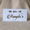 A front-on view of a white acrylic name plate featuring elegant 3D golden "201 Rungta's" lettering and golden leaf accents, resting on a beige fabric surface.