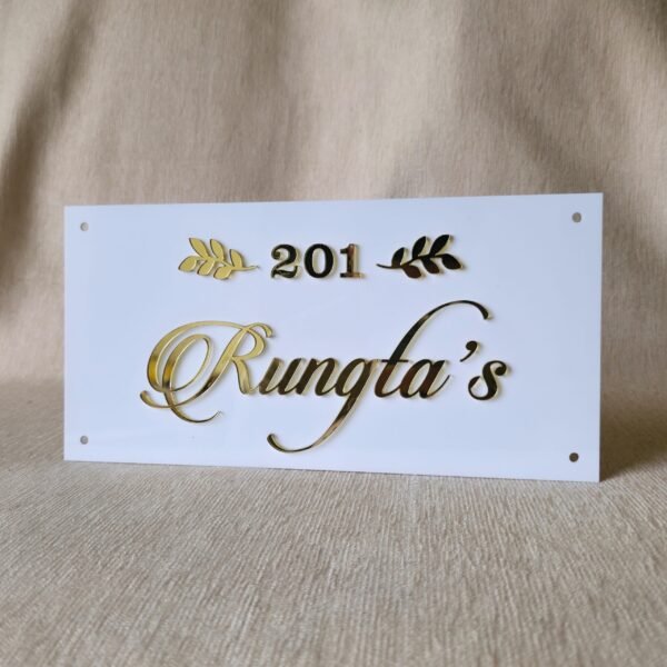 A slightly angled view of a custom white acrylic name plate with raised golden letters "201 Rungta's" and leaf designs, showing the three-dimensional quality of the text.