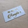 An overhead, close-up shot of a white acrylic name plate with glossy 3D golden text "201 Rungta's" and decorative leaves, highlighting reflections on its surface.