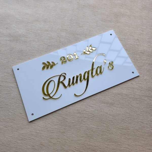An overhead, close-up shot of a white acrylic name plate with glossy 3D golden text "201 Rungta's" and decorative leaves, highlighting reflections on its surface.