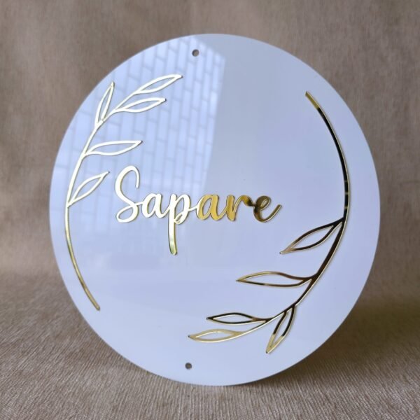 Angled view of a round white acrylic name plate with 'Sapare' and a floral border in 3D gold lettering against a beige backdrop.