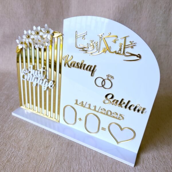 An angled top-down view of the custom white and gold acrylic engagement thumb board, highlighting the calligraphy and names.